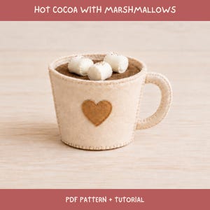 以下が含まれることがあります： 前面に茶色のハートのアップリケが付いたベージュのフェルト製マグカップ。中には茶色のフェルトのココアと3つの白いフェルトのマシュマロが入っています。上部と下部の縁には「HOT COCOA WITH MARSHMALLOWS」および「PDF PATTERN + TUTORIAL」という文字が印刷されています。
