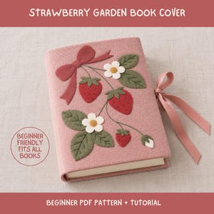 May include: A dusty pink felt book cover featuring an appliqué design of red strawberries, green leaves, and white flowers with yellow centres. A pink ribbon bow is attached to the top left, and a pink ribbon tie extends from the right side.