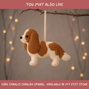 May include: A felt ornament shaped like a Cavalier King Charles Spaniel with cream and brown patches, a black nose, and a black eye. The dog figure hangs from a light beige ribbon attached to a branch. The text reads YOU MAY ALSO LIKE.
