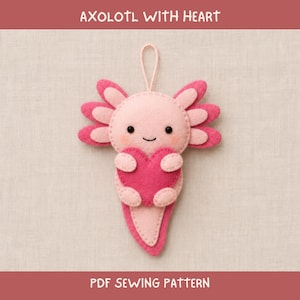 May include: A felt axolotl ornament with a light pink body, darker pink gills, and a bright pink heart held in its arms. The creature has black bead eyes, a small stitched smile, and a loop at the top. Text reads AXOLOTL WITH HEART PDF SEWING PATTERN.
