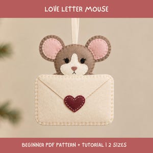 May include: A felt hanging ornament shaped like a brown mouse with pink inner ears holding a cream-coloured envelope. The envelope features a dark red heart appliqué in the centre. The text LOVE LETTER MOUSE and BEGINNER PDF PATTERN + TUTORIAL | 2 SIZES is visible.