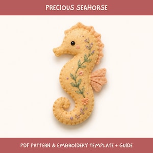 Peut inclure: Une décoration en feutrine couleur pêche en forme d'hippocampe, ornée de broderies de vignes vertes, de petites fleurs roses et de touches violettes. Il possède un œil en perle noire, une nageoire texturée et des coutures visibles. Texte : PRECIOUS SEAHORSE.