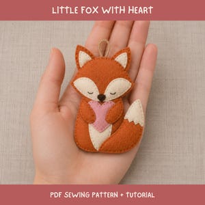 Pode incluir: Um ornamento de feltro em forma de raposa com um pequeno laço no topo, feito de tecido laranja e creme, com um coração rosa nas patas e traços faciais bordados. O texto LITTLE FOX WITH HEART e PDF SEWING PATTERN + TUTORIAL é visível.