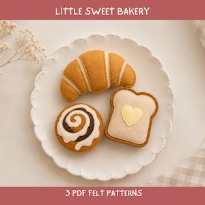 May include: Three felt food items on a white scalloped plate: a golden-brown croissant, a round cinnamon roll with white icing and dark brown swirls, and a piece of toast with a light yellow heart shape. Text reads LITTLE SWEET BAKERY 3 PDF FELT PATTERNS.