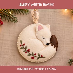 May include: A felt ornament shaped like a curled-up white ermine with a brown-tipped tail, pink inner ears, and a black nose. The body features embroidered green leaves and red berries. A beige ribbon loop is attached for hanging. Text reads SLEEPY WINTER ERMINE BEGINNER PDF PATTERN | 3 SIZES.