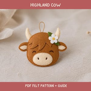 Puede incluir: Un adorno de fieltro marrón con forma de vaca Highland, que presenta un hocico color crema, dos cuernos y una flor blanca con centro amarillo en un lado. Incluye un lazo para colgar. El texto dice HIGHLAND COW PDF FELT PATTERN + GUIDE.