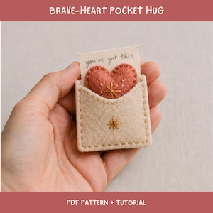 May include: A beige felt pocket containing a small, textured, coral-coloured heart with embroidered star details. The pocket features a matching embroidered star on the front. A small card tucked inside the pocket displays the text you've got this.