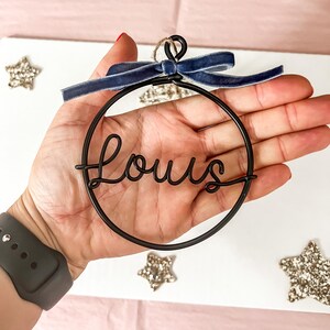K&ouml;nnte beinhalten: Ein schwarzer Drahtschmuck mit dem Namen "Louis" in Schreibschrift. Der runde Schmuck hat eine dunkelblaue Samtschleife oben. Glitzernde Sternverzierungen sind um den Schmuck herum verstreut.