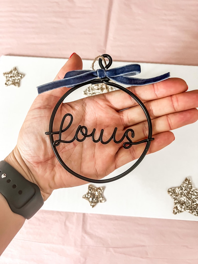 K&ouml;nnte beinhalten: Ein schwarzer Drahtschmuck mit dem Namen "Louis" in Schreibschrift. Der runde Schmuck hat eine dunkelblaue Samtschleife oben. Glitzernde Sternverzierungen sind um den Schmuck herum verstreut.