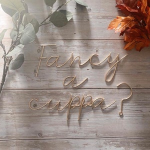 May include: Gold wire letters spelling "Fancy a cuppa?" on a light wood background.