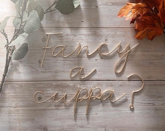May include: Gold wire letters spelling "Fancy a cuppa?" on a light wood background.