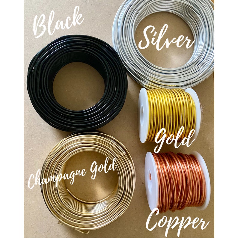 K&ouml;nnte beinhalten: Drahtspulen in Schwarz, Silber, Gold, Champagner-Gold und Kupfer. Die Farbbezeichnungen sind in Schreibschrift &uuml;ber jeder Spule geschrieben. Der Draht ist eng gewickelt.