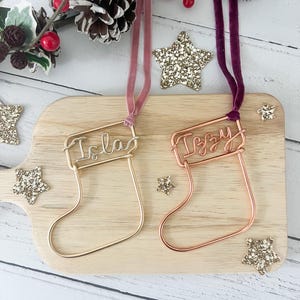 May include: Two gold wire Christmas stocking ornaments with the names "Isla" and "Izzy" written on them. The ornaments are hanging from pink and burgundy velvet ribbon.