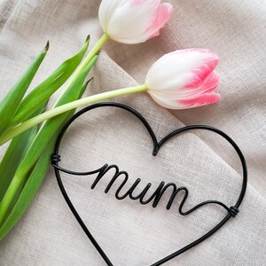 Può includere: Due tulipani rosa e bianchi con steli e foglie verdi poggiano su una superficie di lino beige. Un cuore di filo nero con la parola "mum" in corsivo è posizionato vicino ai fiori, creando una composizione floreale semplice ed elegante.