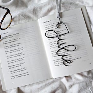 Op de afbeelding: Een zwarte draad bladwijzer in de vorm van een hart met een swirl-ontwerp. De bladwijzer ligt op een open boek met tekst over dansen. De tekst op de pagina luidt "She'd Dance" en "She'd foxtrot and the pots and pans".