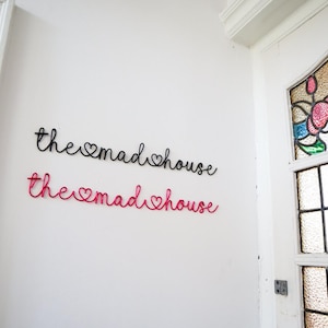 Puede incluir: Dos adornos de pared de alambre con la frase "the mad house" en cursiva, en negro y rosa. Están fijados a una pared blanca. La versión rosa está debajo de la negra. En el fondo, se ve una puerta blanca con vidrieras.