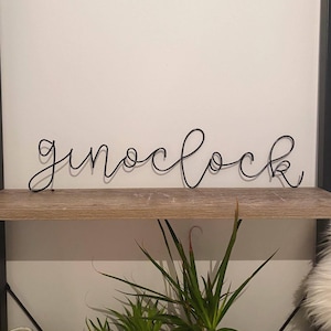 May include: A black metal wire sculpture of the word "o'clock" hanging on a wooden shelf.