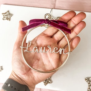 K&ouml;nnte beinhalten: Goldfarbener Drahtschmuck mit dem Namen "Lauren" in Schreibschrift. Der kreisf&ouml;rmige Schmuck hat eine tiefmagentafarbene Samtschleife. Der Hintergrund ist wei&szlig; mit sternf&ouml;rmigen Dekorationen.