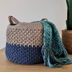 May include: A crocheted storage basket in beige and blue colours. The basket has a handle and is filled with a teal crocheted blanket.