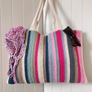 Puede incluir: Un bolso de mano de crochet a rayas con rayas rosas, azules, verdes y beige. El bolso tiene un forro de tela floral y está colgado en una puerta blanca.