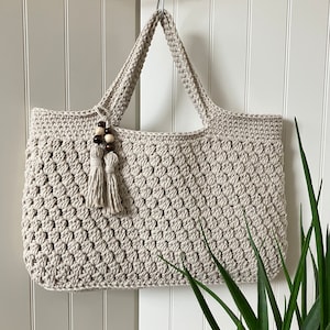 Puede incluir: Un bolso tote de crochet beige con un borla de cuentas marrones y blancas colgando del asa.