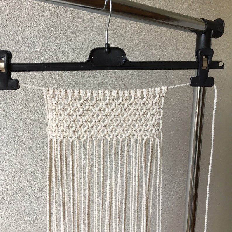 K&ouml;nnte beinhalten: Ein wei&szlig;er Makramee-Wandbehang mit geometrischem Muster und langen Fransen. Der Behang wird an einem Metallb&uuml;gel pr&auml;sentiert.