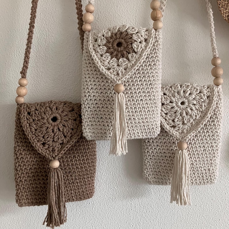 Puede incluir: Tres bolsos de crochet hechos a mano con bandolera en tonos marr&oacute;n y beige. Cada bolso presenta un dise&ntilde;o floral en la solapa, una correa de cuentas de madera y una borla. Los bolsos se exhiben sobre un fondo neutro.