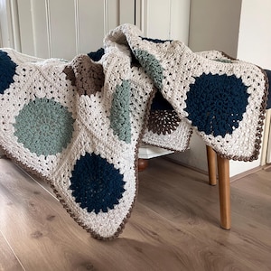 May include: A handmade crochet blanket with a cream-colored base and large, circular patterns in shades of teal, navy, and brown. The blanket is draped over a small stool, showcasing its textured design and intricate stitchwork. The edges are finished with a brown border.