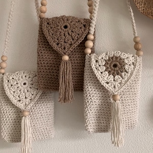 Puede incluir: Tres bolsos de crochet hechos a mano en tonos crema y marr&oacute;n. Cada bolso presenta una solapa decorativa con un dise&ntilde;o floral, una borla y una correa con cuentas de madera. Los bolsos se exhiben sobre un fondo neutro.