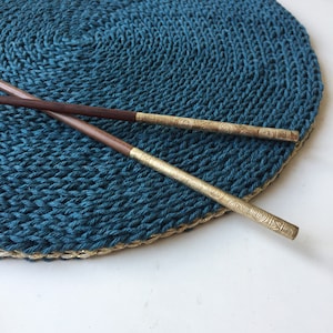 Op de afbeelding: Twee houten eetstokjes met gouden metalen uiteinden liggen op een tealblauwe, ronde, gehaakte placemat met een gouden rand.