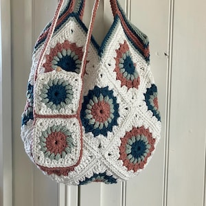 Op de afbeelding: Een handgemaakte gehaakte tote bag opgebouwd uit witte, oudroze en donkergroenblauwe granny squares in een bloemmotief. De tas is voorzien van een bijpassend kleiner tasje aan de schouderband, dat wordt gesloten met een houten kraalknoop.