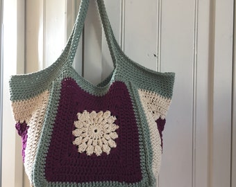 May include: A crocheted tote bag with a green, white, and purple color scheme. The bag has a large, white crocheted flower in the center.