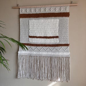 May include: A handcrafted wall hanging in neutral tones. The piece features a light grey crocheted background with brown and white textured details. The bottom edge has long, white fringe, and it hangs from a wooden dowel.