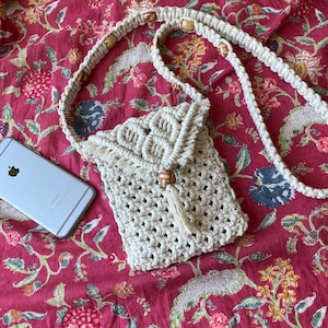 Op de afbeelding: Een witte gehaakte schoudertas met een bruine houten kralenaccent en een kwast. De tas ligt op een rode bloemachtige stoffen achtergrond.