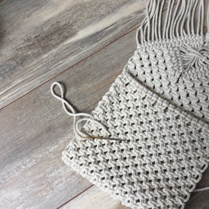 K&ouml;nnte beinhalten: Nahaufnahme einer wei&szlig;en Makramee-Tasche vor einem Holz-Hintergrund. Die Tasche ist aus dickem Seil gefertigt und hat einen Fransen-Detail. Eine Nadel ist in der Tasche sichtbar.