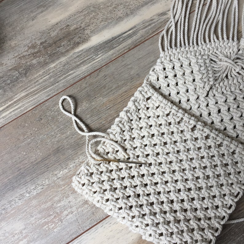 K&ouml;nnte beinhalten: Nahaufnahme einer wei&szlig;en Makramee-Tasche vor einem Holz-Hintergrund. Die Tasche ist aus dickem Seil gefertigt und hat einen Fransen-Detail. Eine Nadel ist in der Tasche sichtbar.
