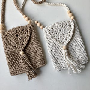 Puede incluir: Dos bolsos cruzados de ganchillo, uno beige y otro blanco roto. Cada uno tiene una solapa con un dise&ntilde;o floral, una borla y una correa con cuentas de madera. Los bolsos rectangulares parecen hechos a mano.