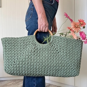 Könnte beinhalten: Eine große, salbeigrüne Häkeltasche mit einem kreisförmigen Holzgriff. Die Tasche ist mit rosa und orangefarbenen Blumen gefüllt. Die Tasche wird von einer Person gehalten, die eine blaue Jeans trägt.