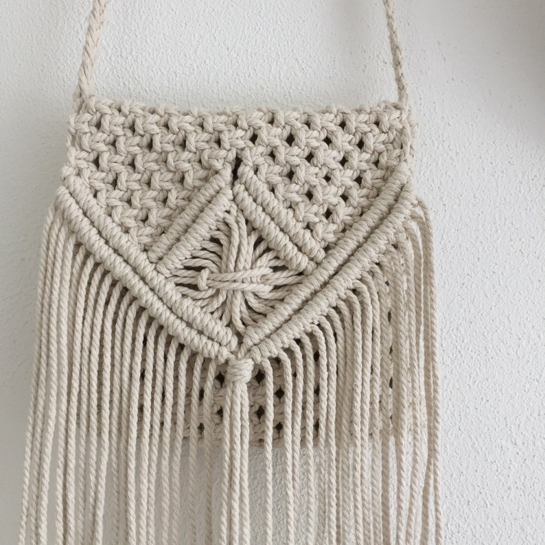 K&ouml;nnte beinhalten: Eine beige Makramee-Tasche mit Fransenrand. Die Tasche hat ein geometrisches Muster und ist aus Naturfasern gefertigt.