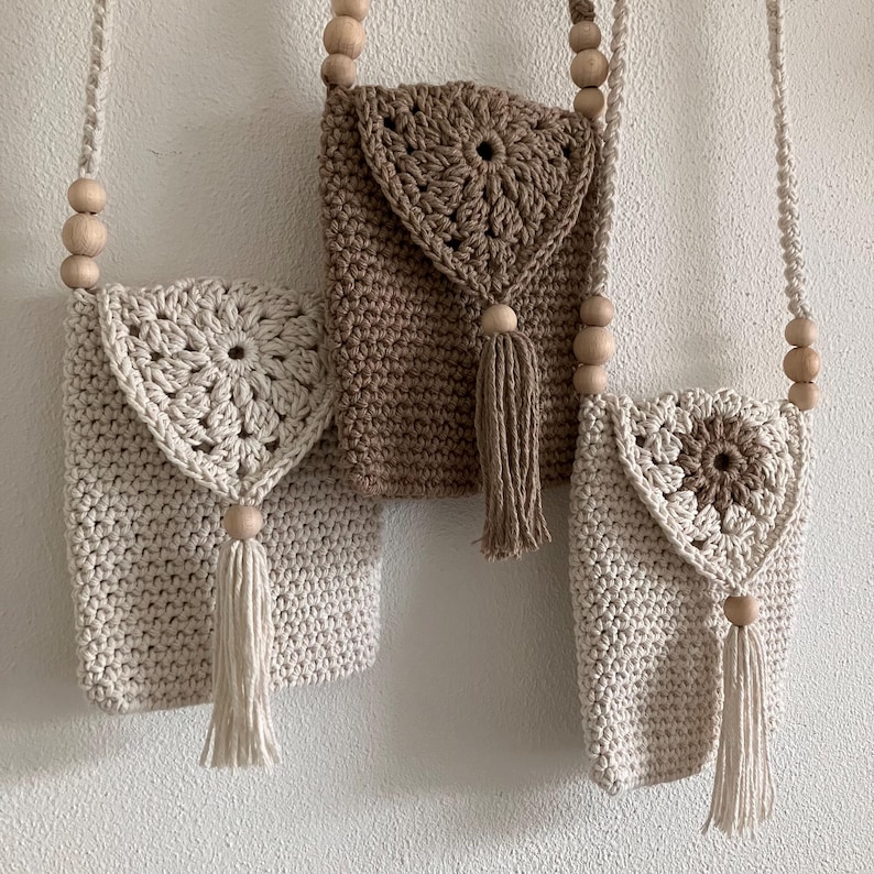 Puede incluir: Tres bolsos de crochet hechos a mano en crema y marr&oacute;n. Cada bolso presenta un dise&ntilde;o floral decorativo en la solapa, una correa de cuentas de madera y una borla. Los bolsos cuelgan contra una pared blanca.