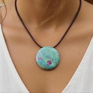 Può includere: Una collana con pendente rotondo con una grande pietra lucida con un motivo screziato turchese e rosa. Il pendente è sospeso a una sottile collana con cordino marrone scuro. La collana è indossata sopra un top bianco.