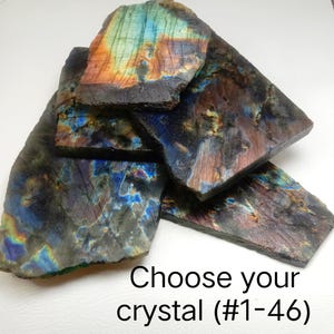 May include: A collection of flat, irregular labradorite crystal slabs featuring iridescent flashes of blue, gold, and orange against a dark gray base. The text "Choose your crystal (#1-46)" is printed in black at the bottom of the image.