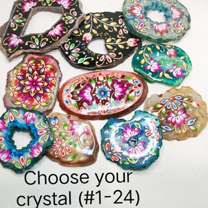 May include: A collection of polished agate slices featuring hand-painted floral designs in vibrant pink, blue, green, and gold colors. Each stone has a unique shape and natural edge. The text at the bottom reads: Choose your crystal (#1-24).