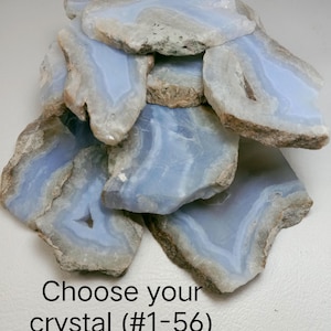 May include: A collection of irregular, flat blue lace agate crystal slices with translucent, banded patterns and rough, natural tan edges. The stones are stacked together, and the text "Choose your crystal (#1-56)" is printed at the bottom.