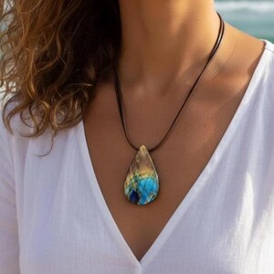 Könnte beinhalten: Halskette mit einem tropfenförmigen Anhänger aus blauem, goldenem und braunem Edelstein. Der Anhänger hängt an einer dünnen, dunkelbraunen Kordel. Die Halskette wird über einem weißen Oberteil getragen.