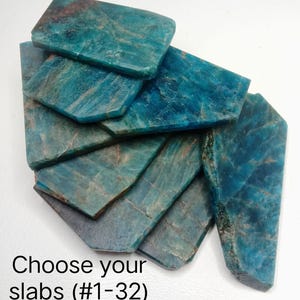 Può includere: Una collezione di lastre minerali di apatite blu, piatte e levigate, con venature naturali e texture cristalline visibili. Le pietre sono disposte in una pila sovrapposta. Il testo "Choose your slabs (#1-32)" è stampato in nero in basso a sinistra.