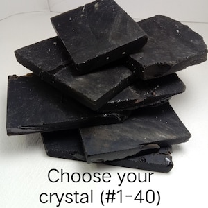 Peut inclure: Une pile de dalles de cristal noir foncé, plates et irrégulières, aux surfaces lisses et aux bords dentelés. Le texte "Choose your crystal (#1-40)" est imprimé en noir au bas de l'image, indiquant une sélection de pièces individuelles.