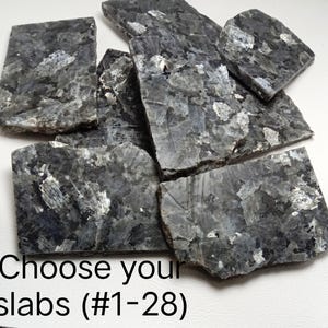 May include: A collection of flat, rectangular labradorite stone slabs featuring a dark grey base with iridescent blue and silver crystalline inclusions. The text "Choose your slabs (#1-28)" is printed in black at the bottom of the image.