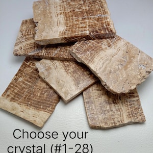 May include: A collection of five rectangular, flat, polished stone slabs featuring natural banded patterns in shades of tan, beige, and brown. The text "Choose your crystal (#1-28)" is printed in black at the bottom of the image.