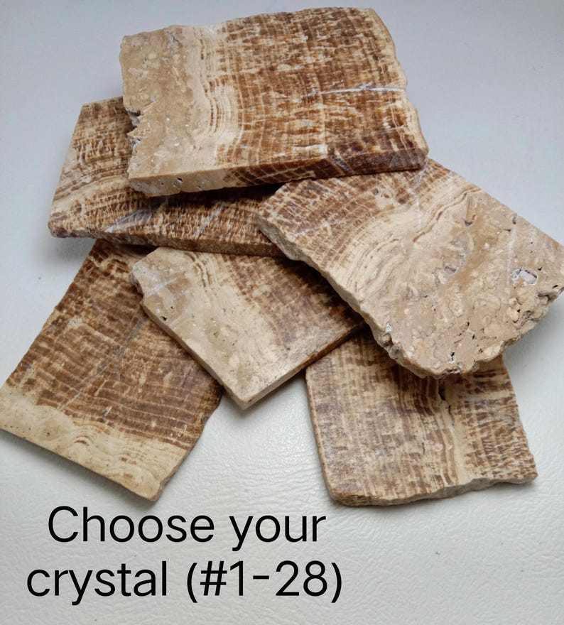 May include: A collection of five rectangular, flat, polished stone slabs featuring natural banded patterns in shades of tan, beige, and brown. The text "Choose your crystal (#1-28)" is printed in black at the bottom of the image.